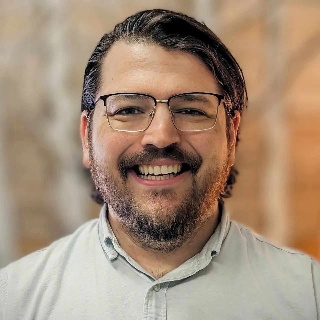 Smiling man with glasses and a light shirt, against a blurred background.